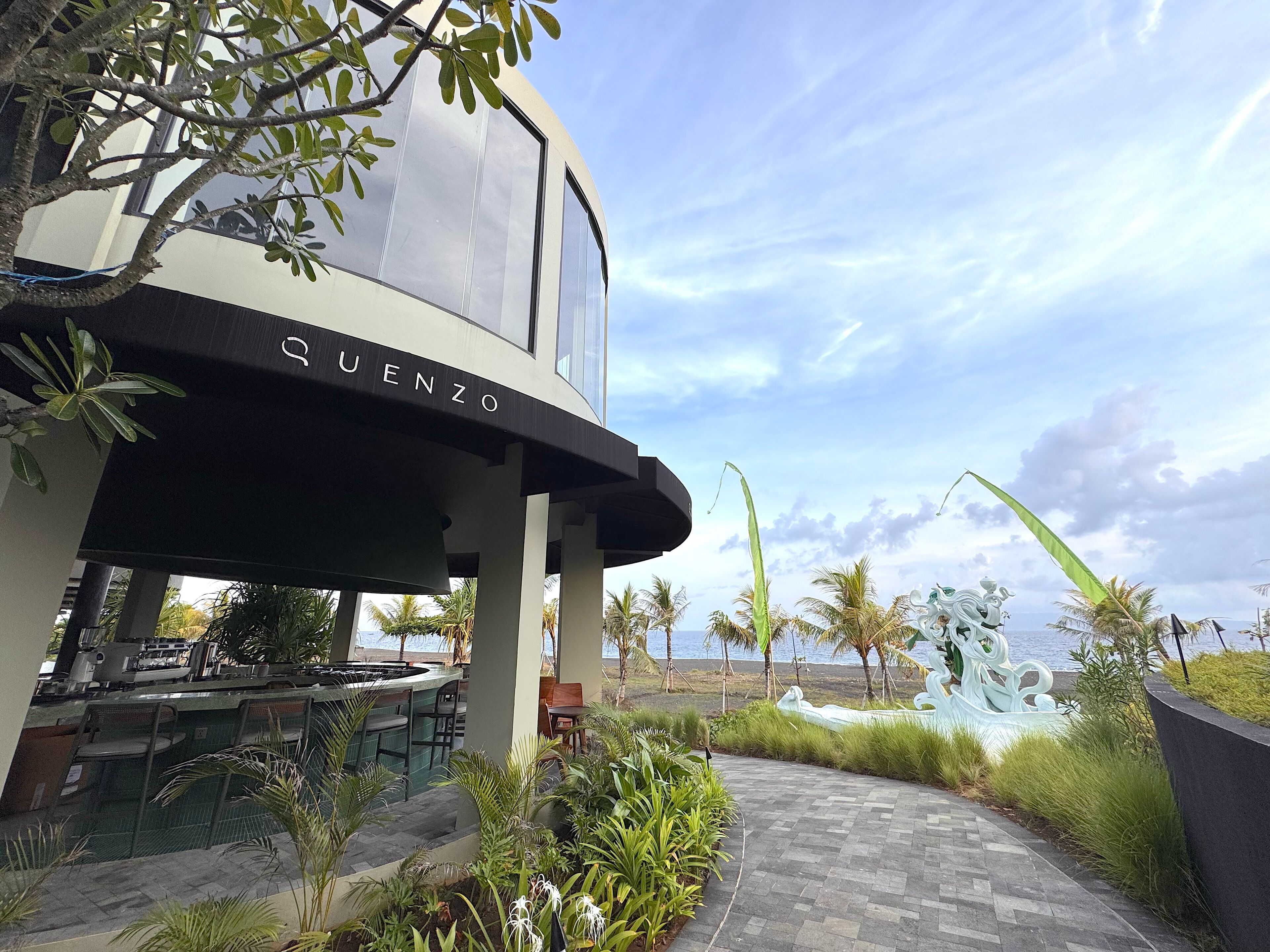 A modern building with the name 'QUENZO' on its facade, surrounded by lush greenery and palm trees, with a view of the sea in the background.