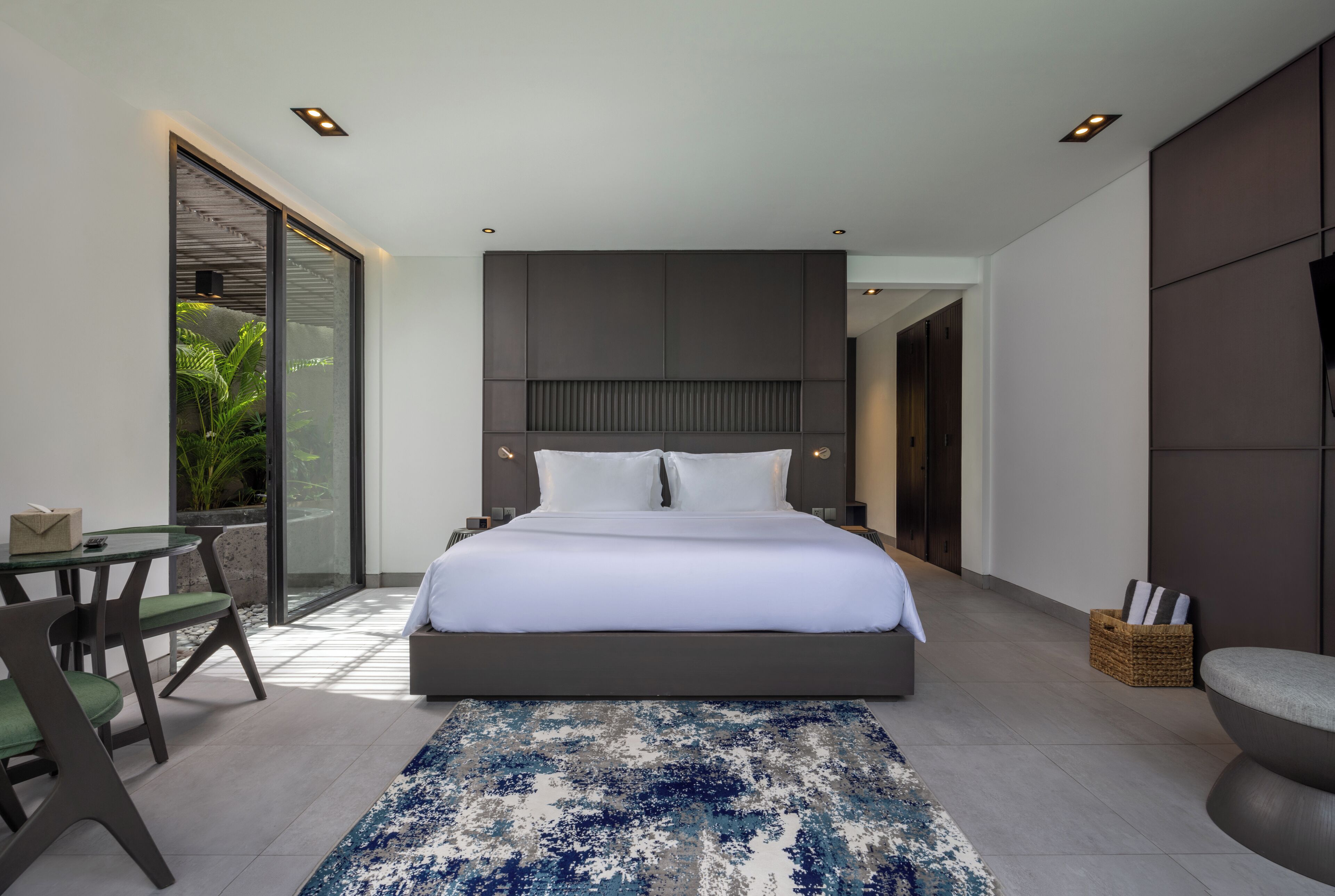 A modern bedroom with a large bed, a glass door leading to a balcony, a small table with chairs, and a large window.