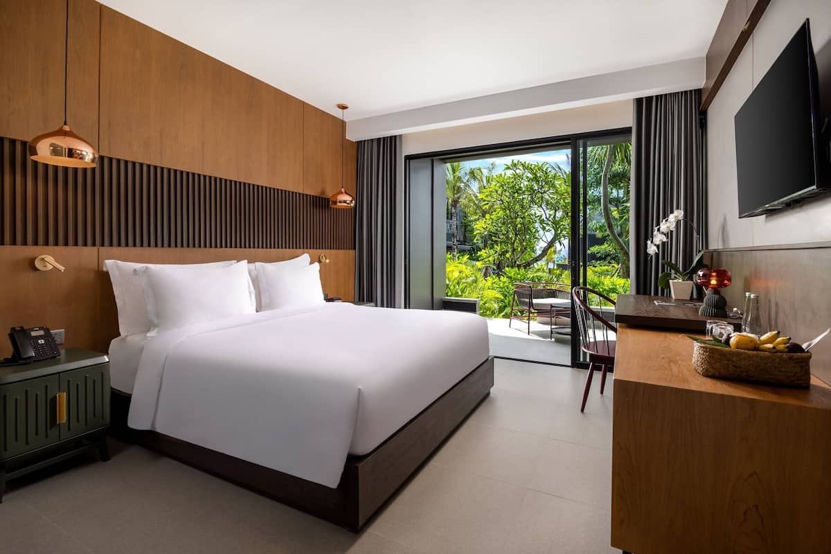 A modern hotel room with a large bed, a desk, a TV, and a balcony with a view of greenery.