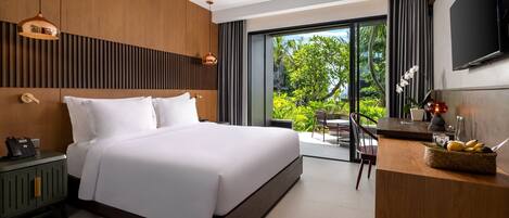 A modern hotel room with a large bed, a desk, a TV, and a balcony with a view of greenery.
