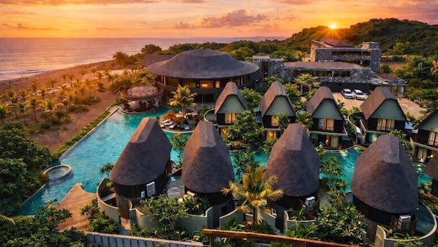 Een resort met een zwembad, bungalows met rieten daken en uitzicht op de oceaan bij zonsondergang.