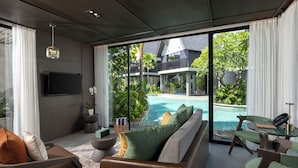 Een moderne woonkamer met een grote glazen deur naar een zwembadgedeelte, een flatscreen-tv aan de muur en een zithoek met kussens en een klein tafeltje.