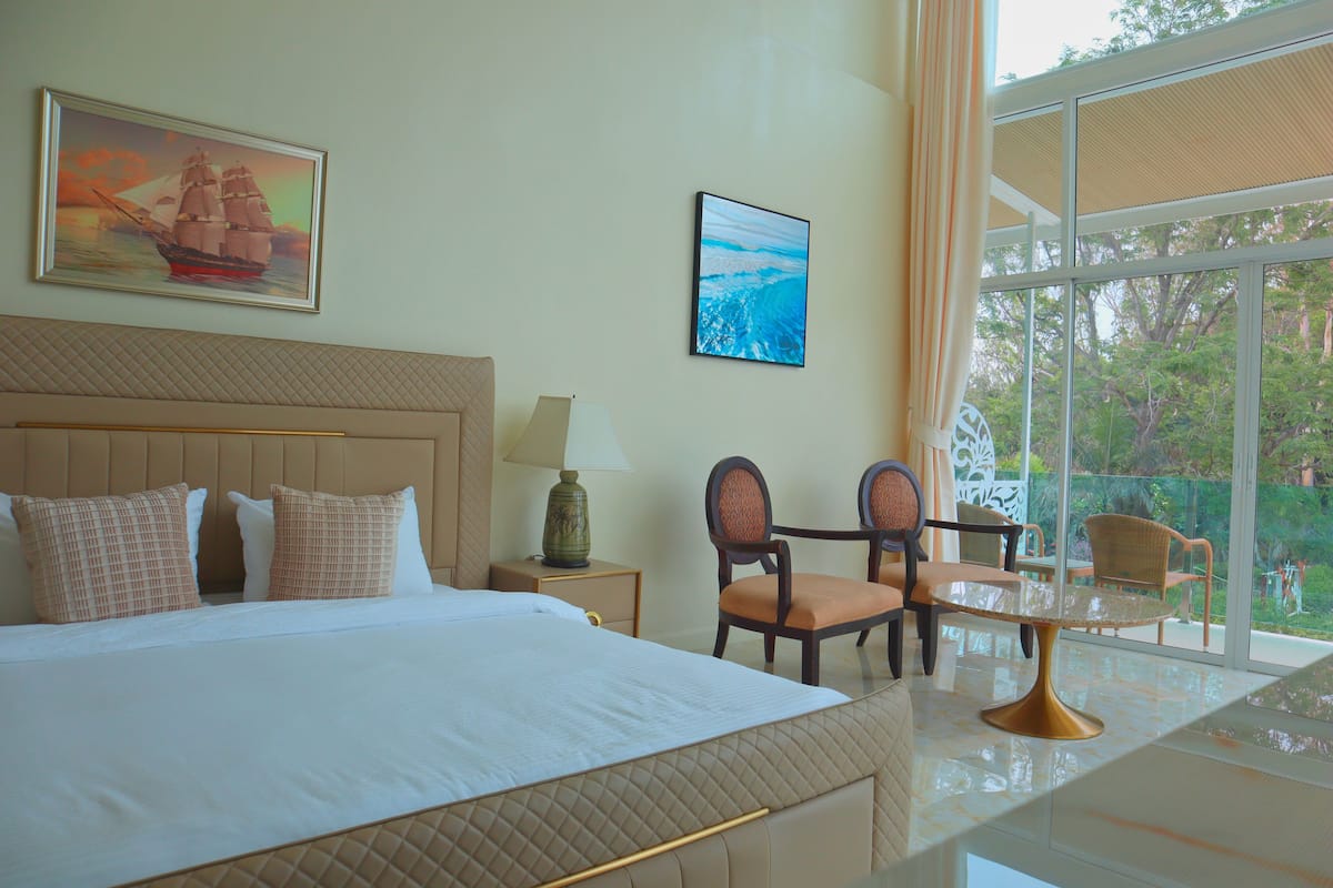 A hotel room with a large bed, a desk with a chair, a window with a view of trees, and two paintings on the wall.