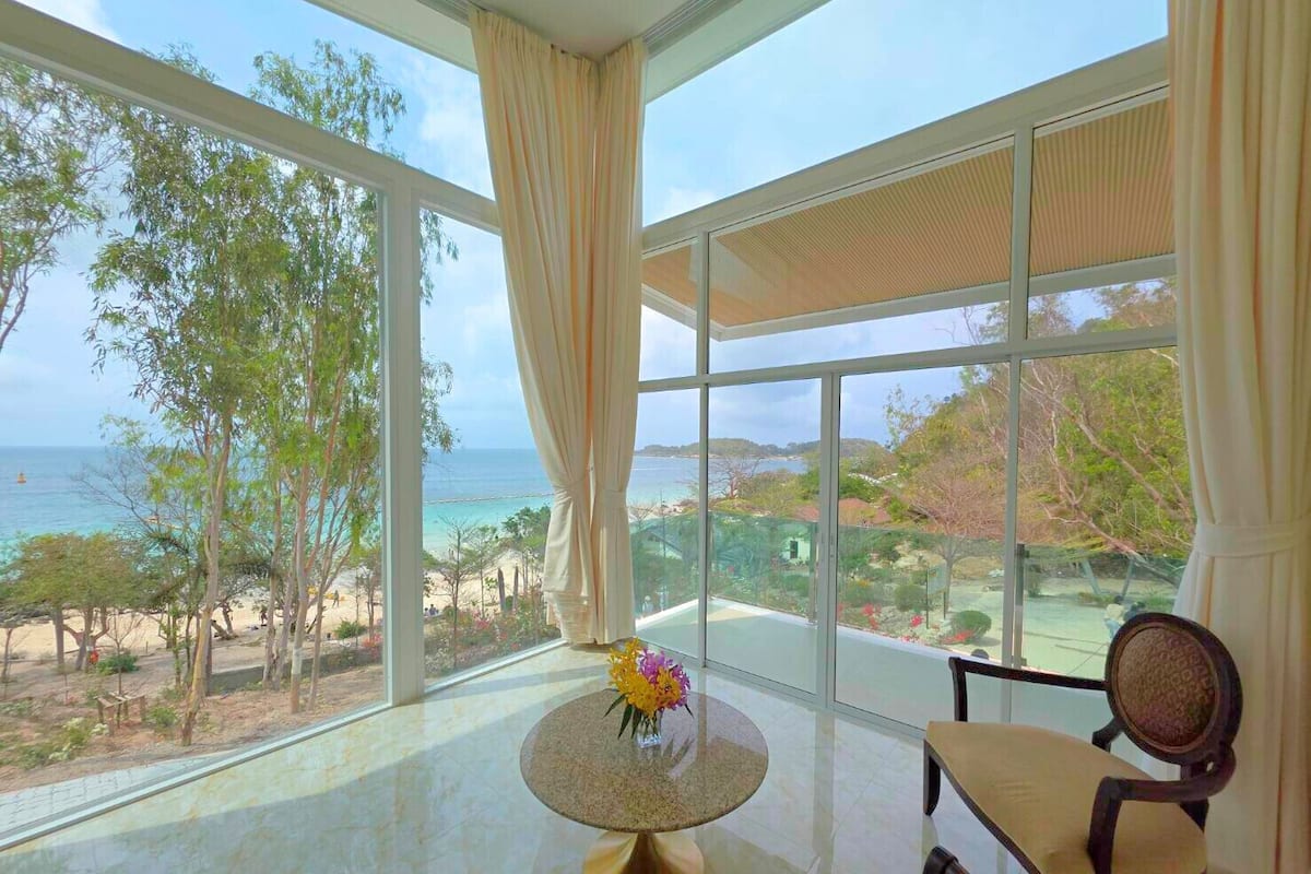 A room with a large window offering a view of the beach and ocean. There is a round table with a vase of flowers and a chair.