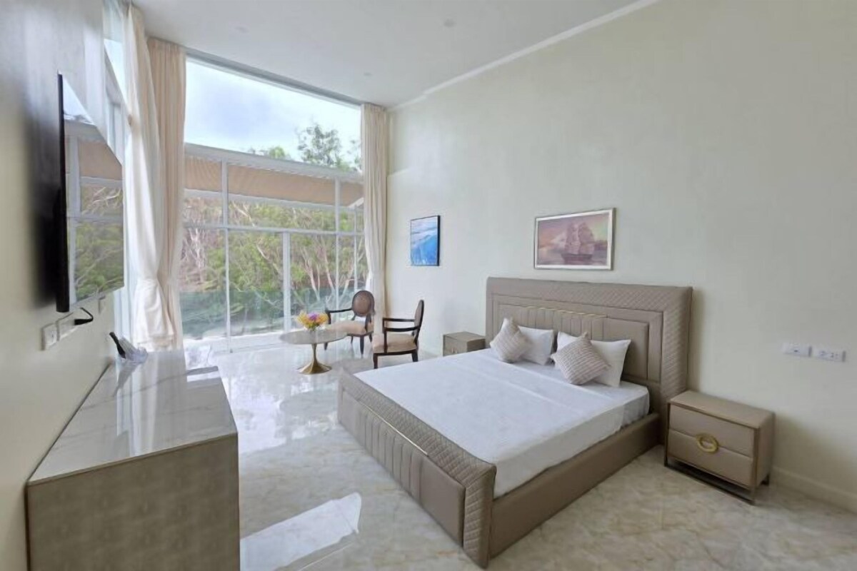 A modern hotel room with a large bed, a desk, and a chair. There is a window with curtains, a TV mounted on the wall, and a small table with a vase of flowers.