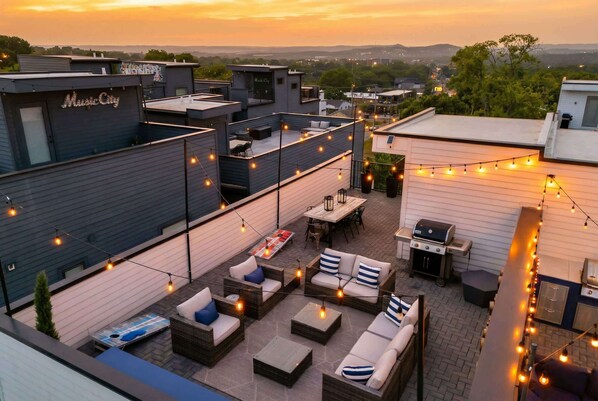 A rooftop patio with outdoor seating, string lights, and a view of the cityscape at sunset.