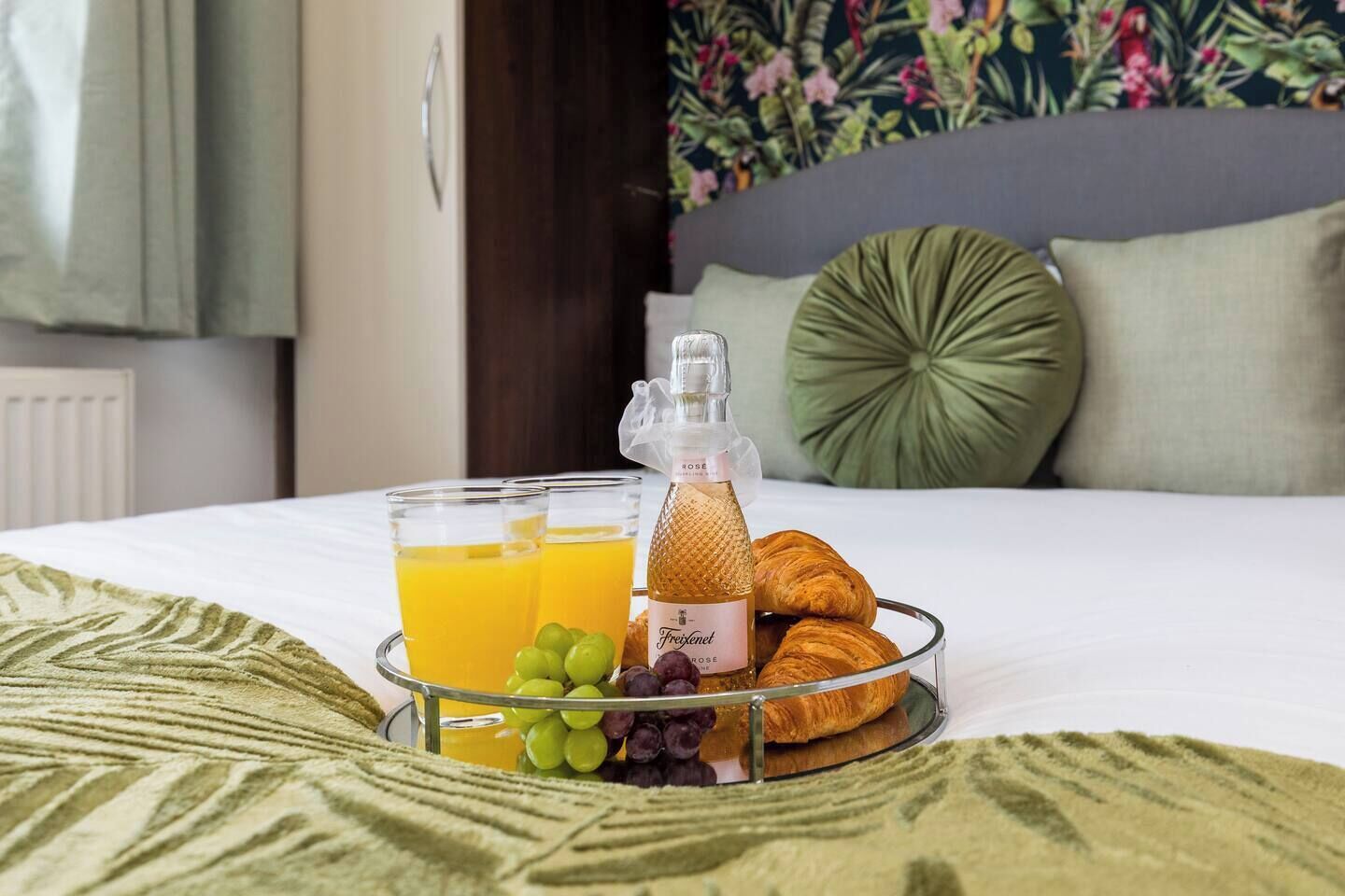 Un lit bien fait, recouvert d’une couvre-lit à motifs verts, un plateau avec des croissants, des raisins et une bouteille de jus de fruits, et un mur décoré de motifs floraux en arrière-plan.