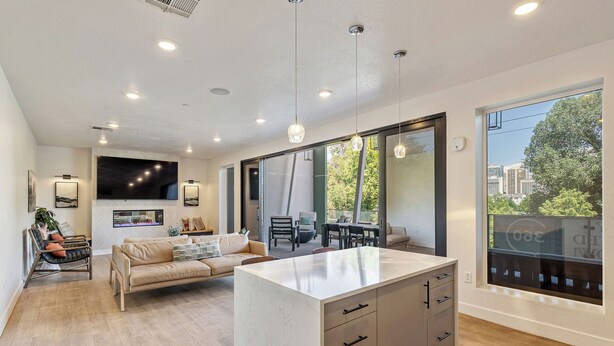 A modern kitchen with a large island, a dining area with a table and chairs, a sofa, and a large window with a city view.