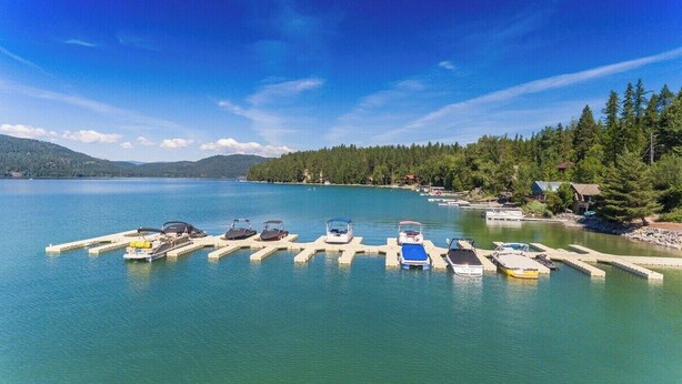 A marina with boats docked at wooden piers, surrounded by a forested shoreline and a calm lake. - Mountain Harbor Whitefish Captains Quarters 151 (Whitefish)