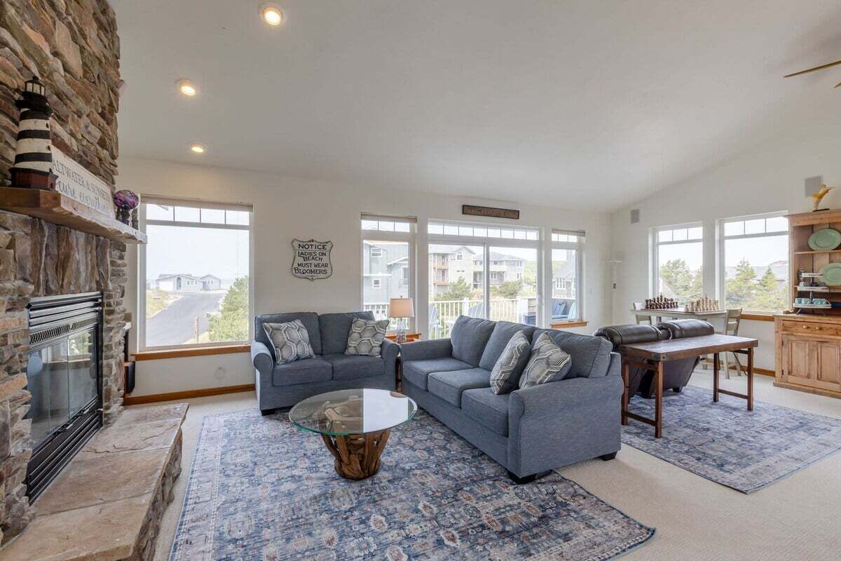 A spacious living room with a stone fireplace, two grey sofas, a glass coffee table, and a view of the neighborhood through large windows.