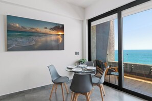 Un espace repas avec vue sur l’océan, un tableau représentant un coucher de soleil et un balcon donnant sur la mer.