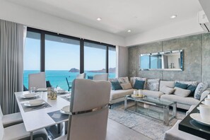 Una sala de estar moderna con sofá, mesa de centro y mesa de comedor lista para una comida. Amplias ventanas ofrecen vistas al océano.