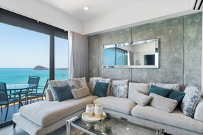 A modern living room with a large window overlooking the sea, a sofa with patterned cushions, a glass coffee table, and a mirror on the wall.