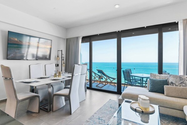 A modern hotel room with a dining area, a sofa, and a large balcony with a view of the ocean.