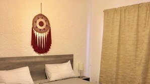 Un lit bien fait, avec du linge de lit blanc et deux serviettes enroulées, un attrape-rêves accroché au mur, un ventilateur de plafond et une fenêtre avec un rideau.