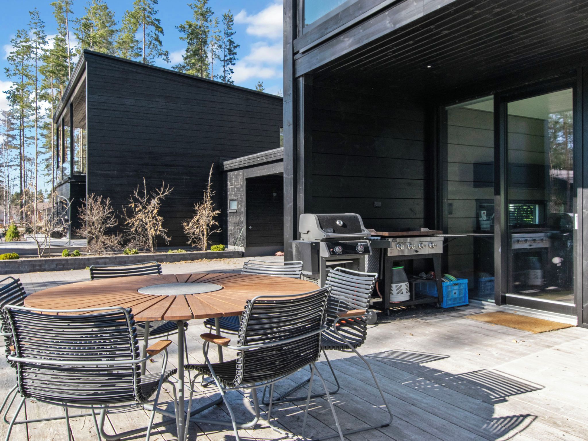 Et udendørs spiseområde med et træbord og metalstole, en grill og en moderne bygning i baggrunden.