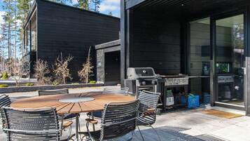 Et udendørs spiseområde med et træbord og metalstole, en grill og en moderne bygning i baggrunden.