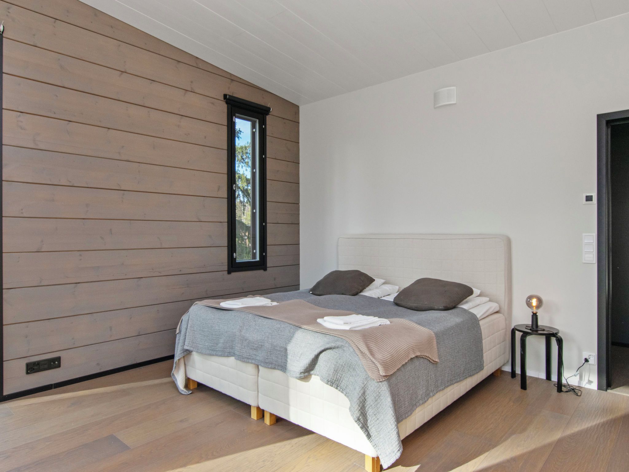 A modern bedroom with a wooden wall, a large bed with a headboard, and a small bedside table with a lamp.
