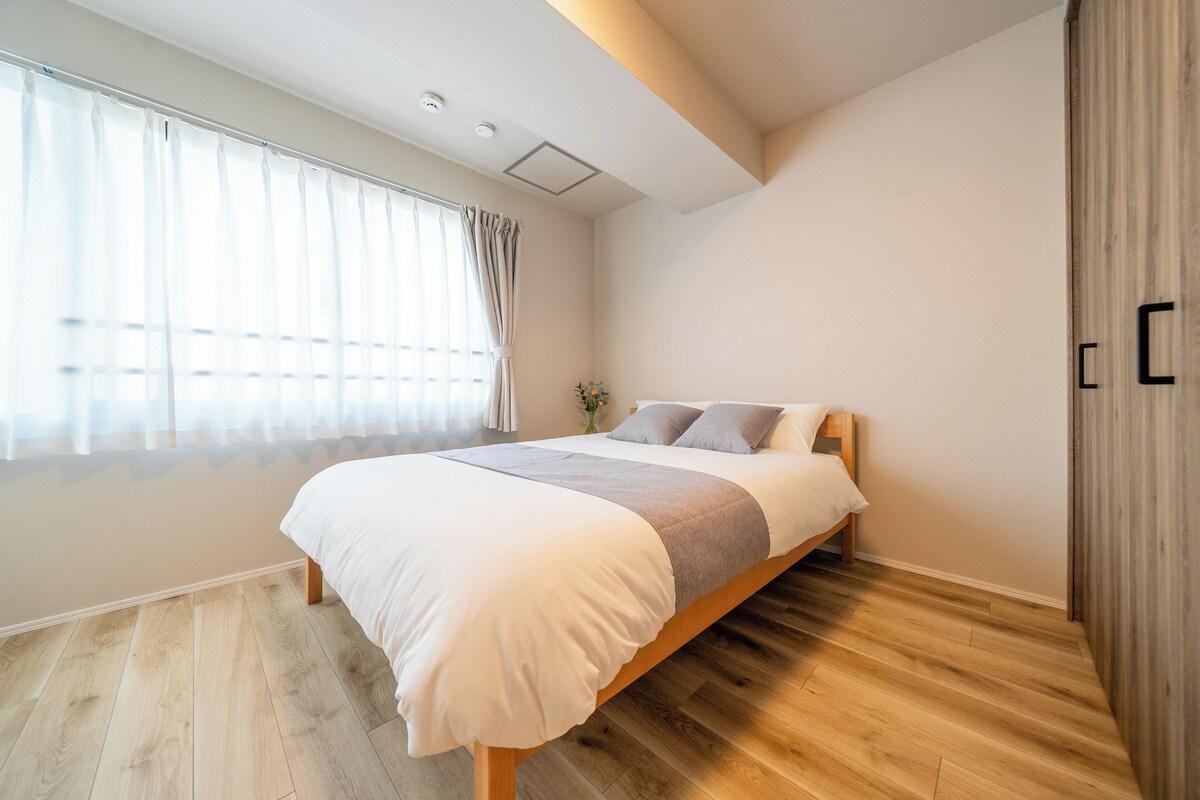 Une chambre moderne avec un plancher en bois, un grand lit recouvert d’une couette blanche et grise, et une fenêtre ornée de rideaux légers.