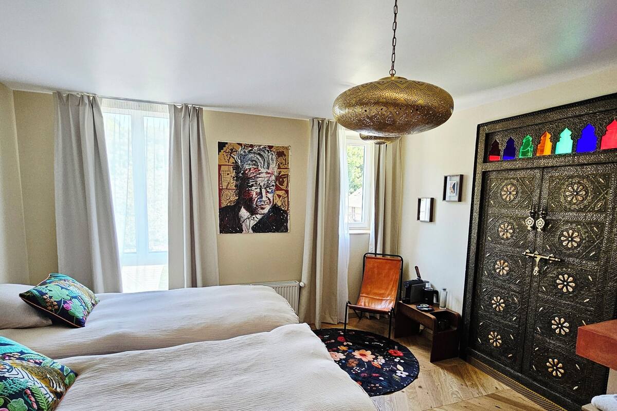 Una habitación con dos camas, una lámpara colgante, una silla, una puerta decorativa y un cuadro en la pared.