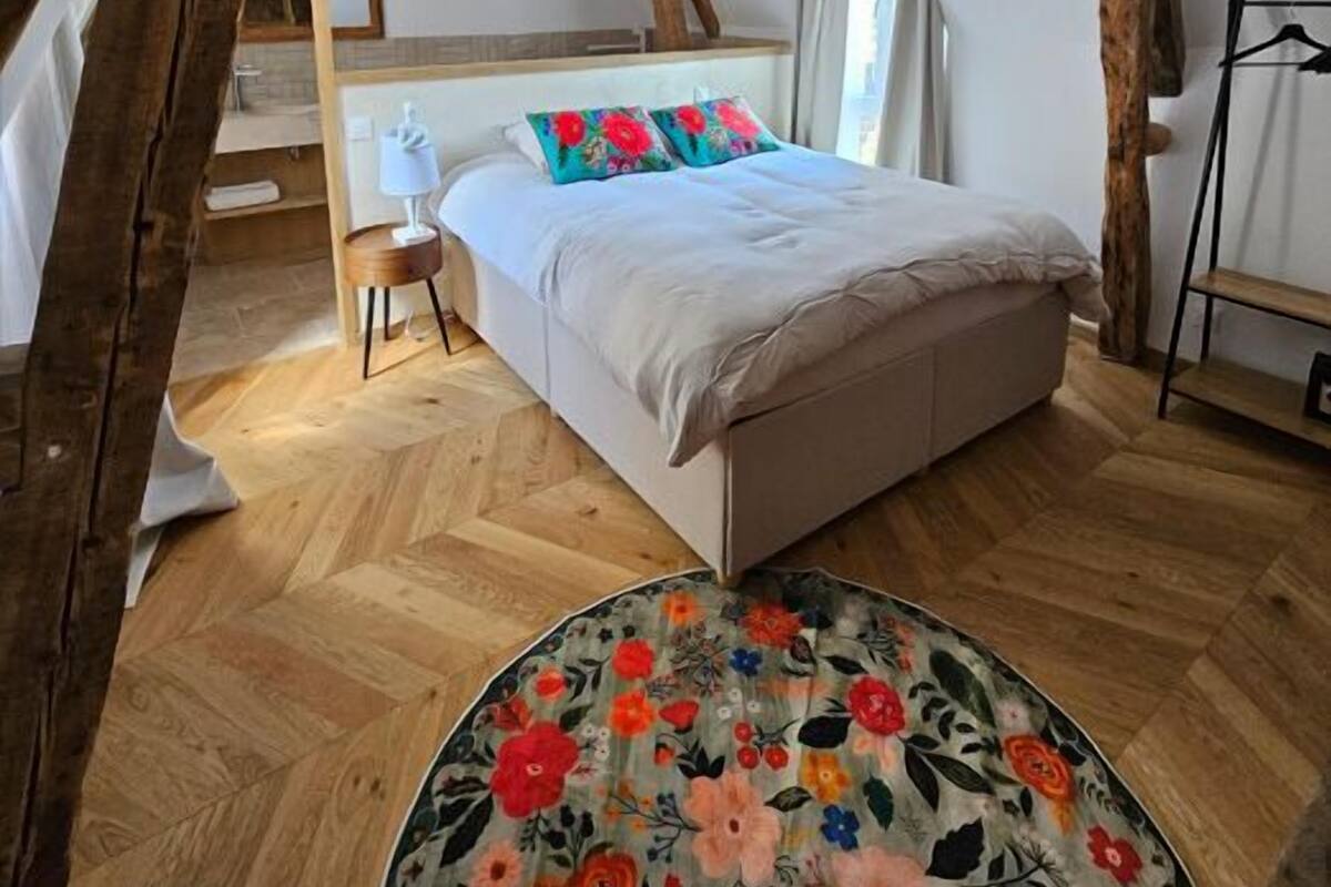 Un dormitorio con techo inclinado, vigas de madera, una cama con almohadas coloridas, una mesita de noche pequeña y una alfombra redonda.