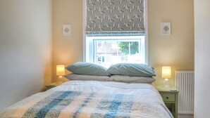 A bedroom with a bed, two bedside lamps, a window with a seagull pattern blind, and a radiator. - You will just love the open plan ground floor of this historic cottage. (Flamborough)