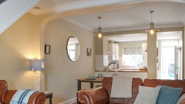 A cozy living room with leather sofas, a wooden coffee table, and a dining table. A kitchen is visible in the background with white cabinets and a window. - You will just love the open plan ground floor of this historic cottage. (Flamborough)