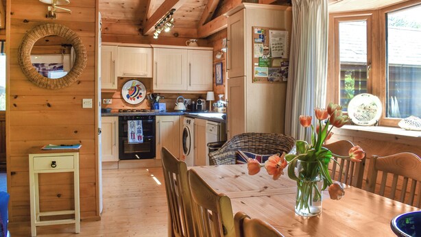A wooden interior with a dining table, chairs, a vase with flowers, a kitchen area with appliances, and a window with curtains.