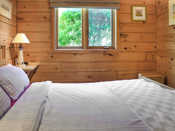 A wooden cabin bedroom with a bed, a nightstand, a lamp, and a window with a view of greenery.