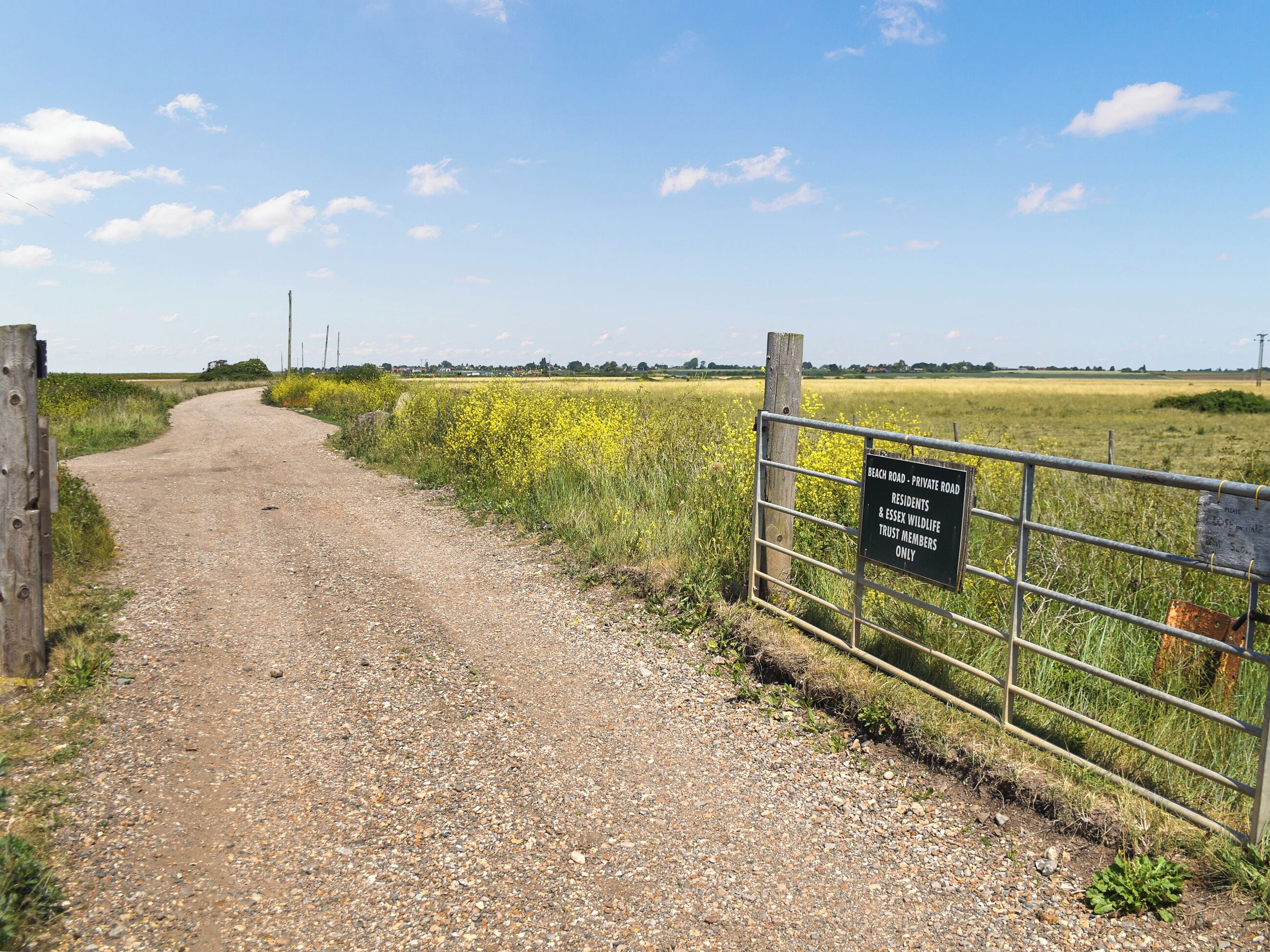 Een grindpad dat door een landelijk gebied loopt, met een metalen poort en een bord waarop staat dat het een privéweg is, alleen toegankelijk voor bewoners en leden van de natuurtuin.