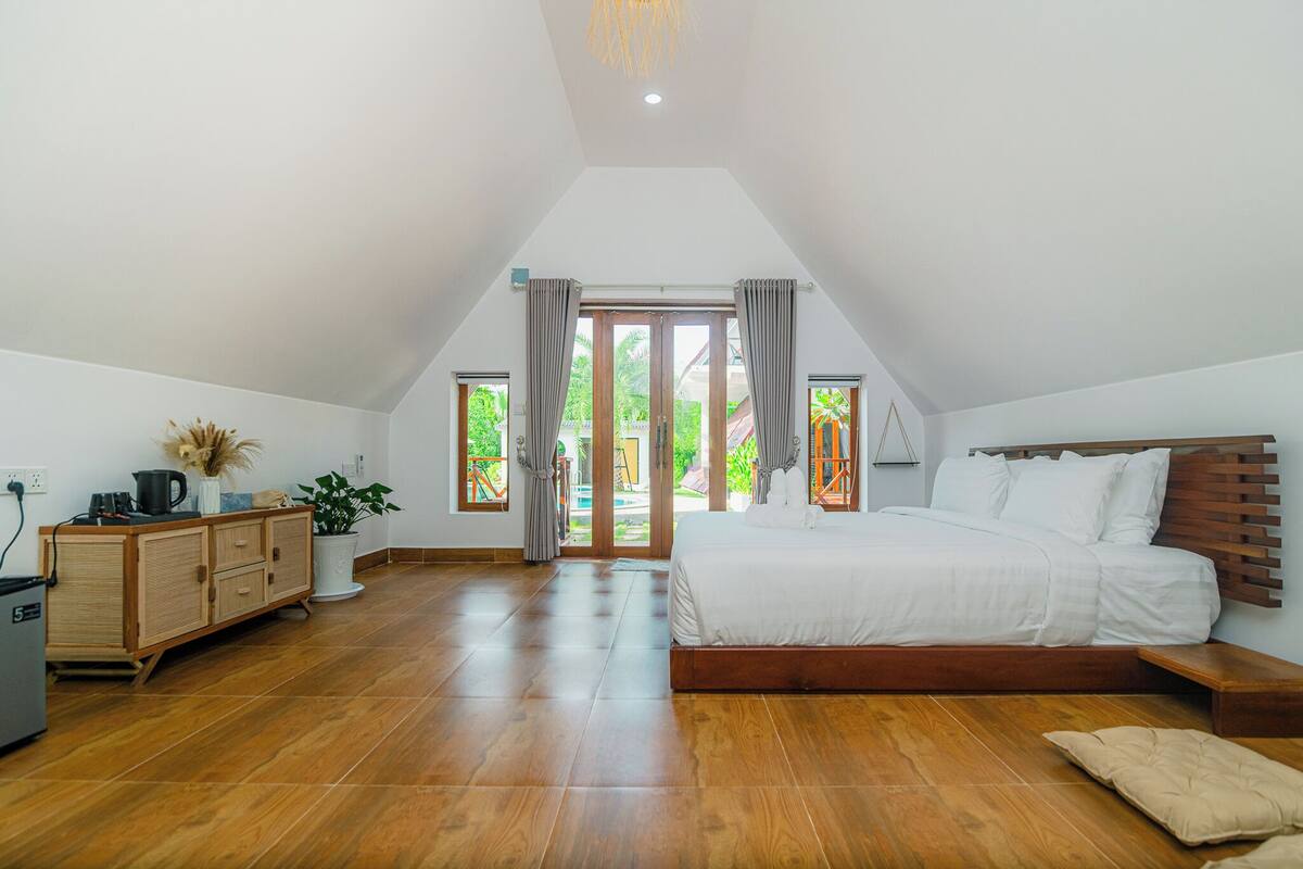 A bedroom with a large bed, wooden floor, a dresser, a plant, and a view through a glass door.