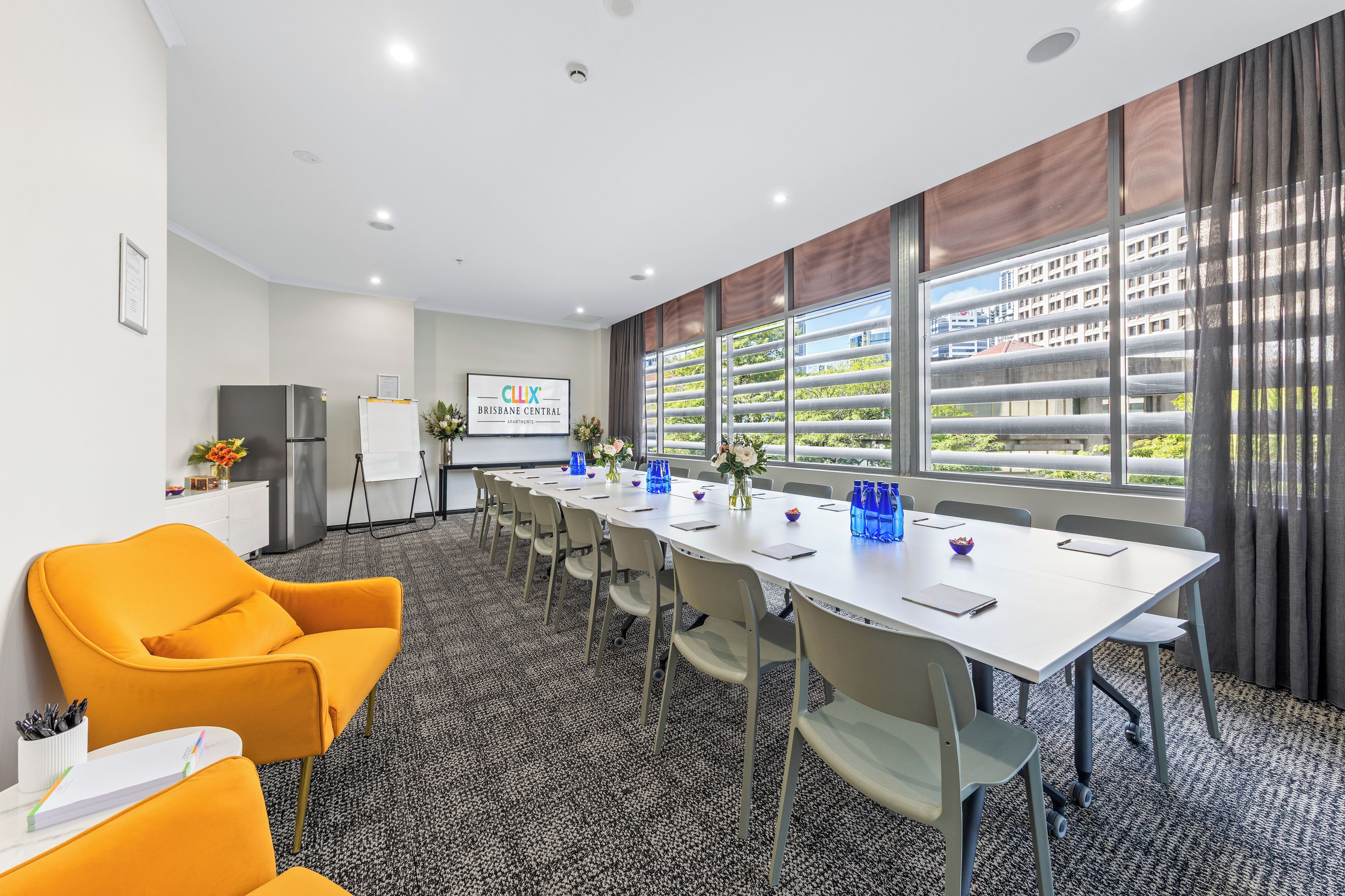 A conference room with a long table, chairs, a projector screen, and a view of buildings through large windows.