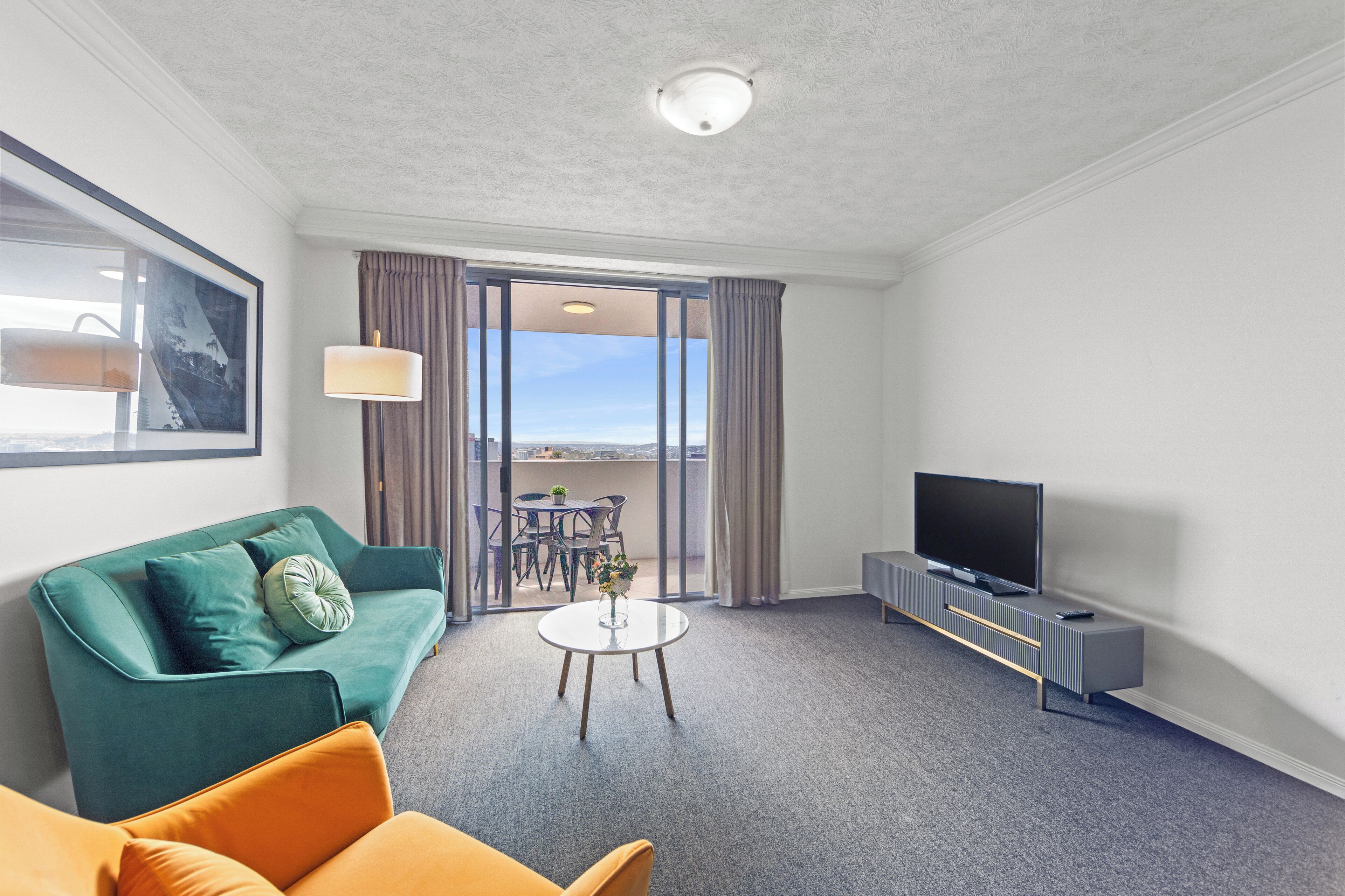 A modern hotel room with a green sofa, a smaller orange sofa, a flat-screen TV, a small round coffee table, and a balcony with a table and chairs.