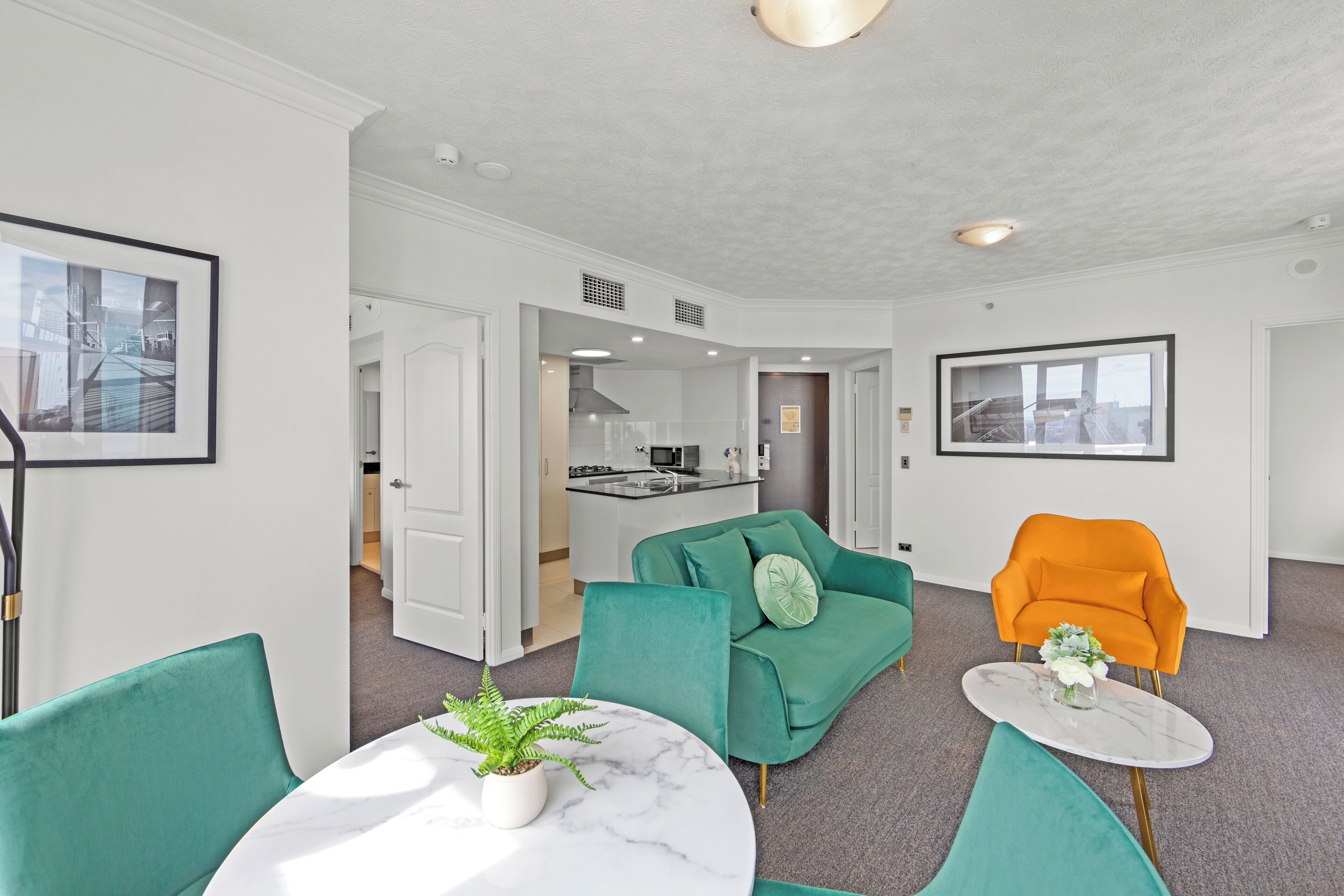 Un salon moderne avec des fauteuils turquoise et orange, une table en marbre et une cuisine en arrière-plan.