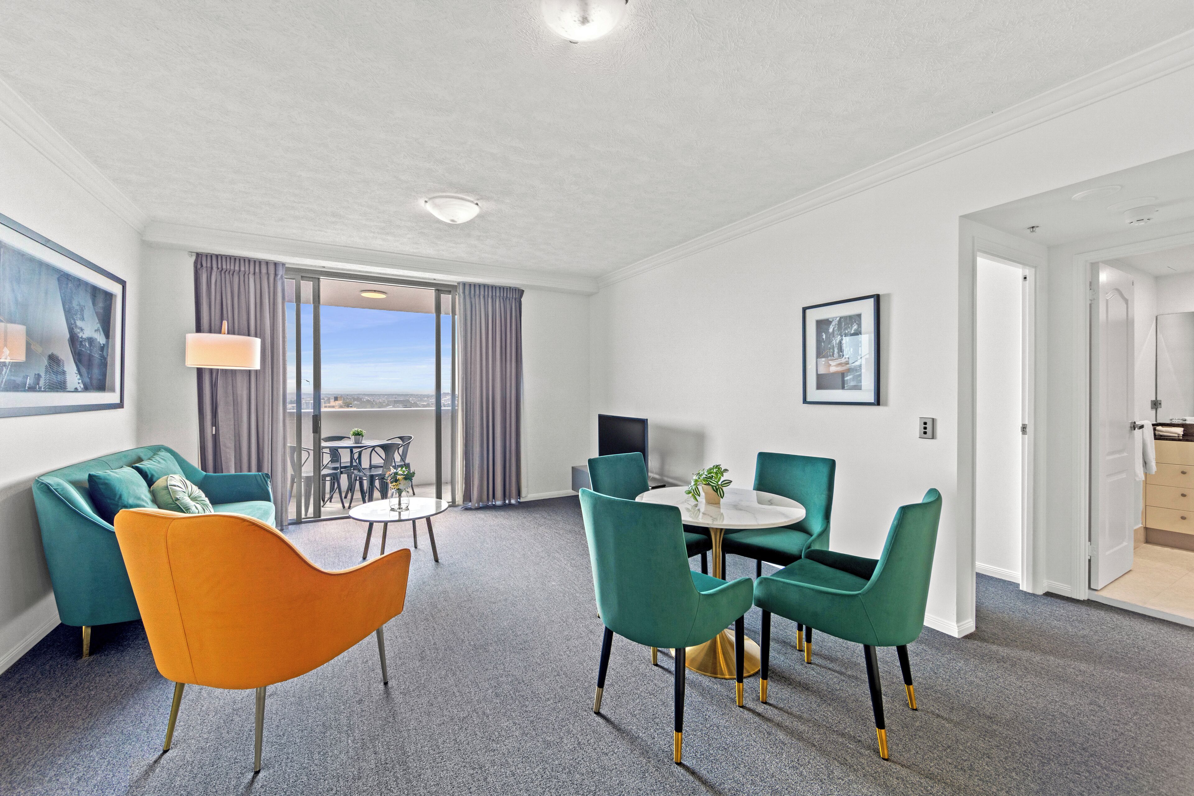 A modern hotel room with a sofa, a dining table with chairs, a TV, and a balcony with curtains.