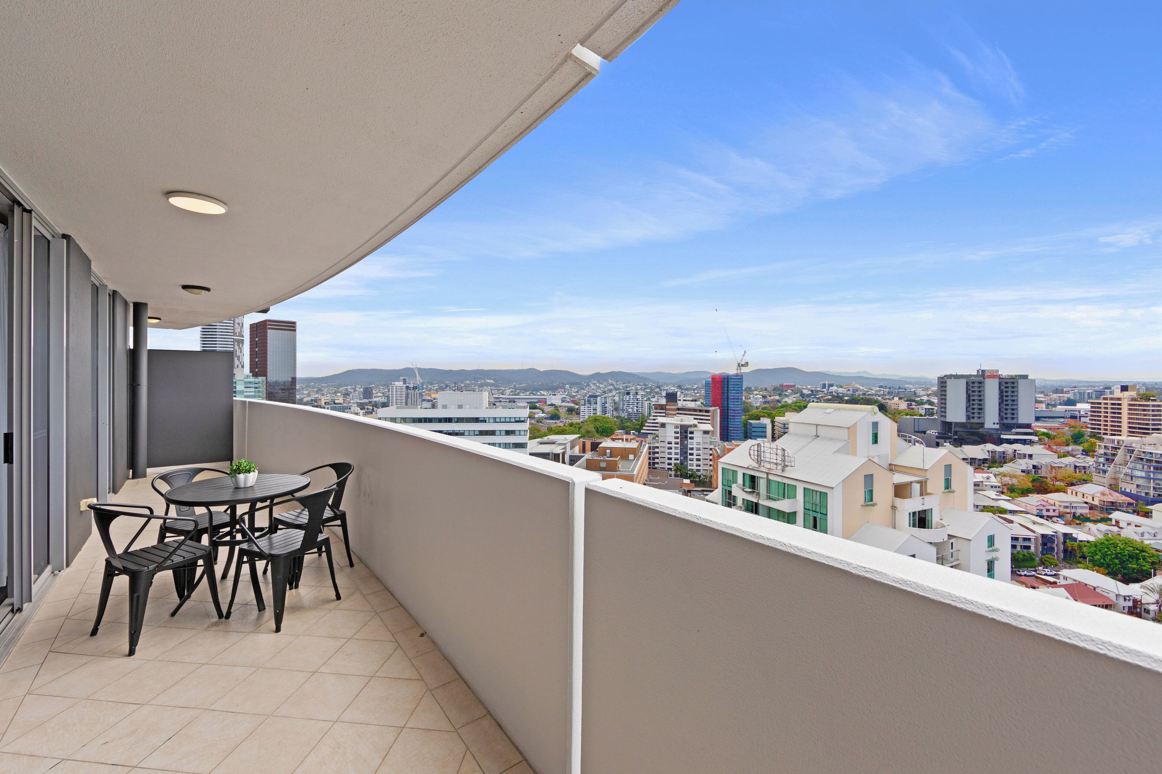 A balcony with a table and chairs, overlooking a cityscape with buildings and mountains.