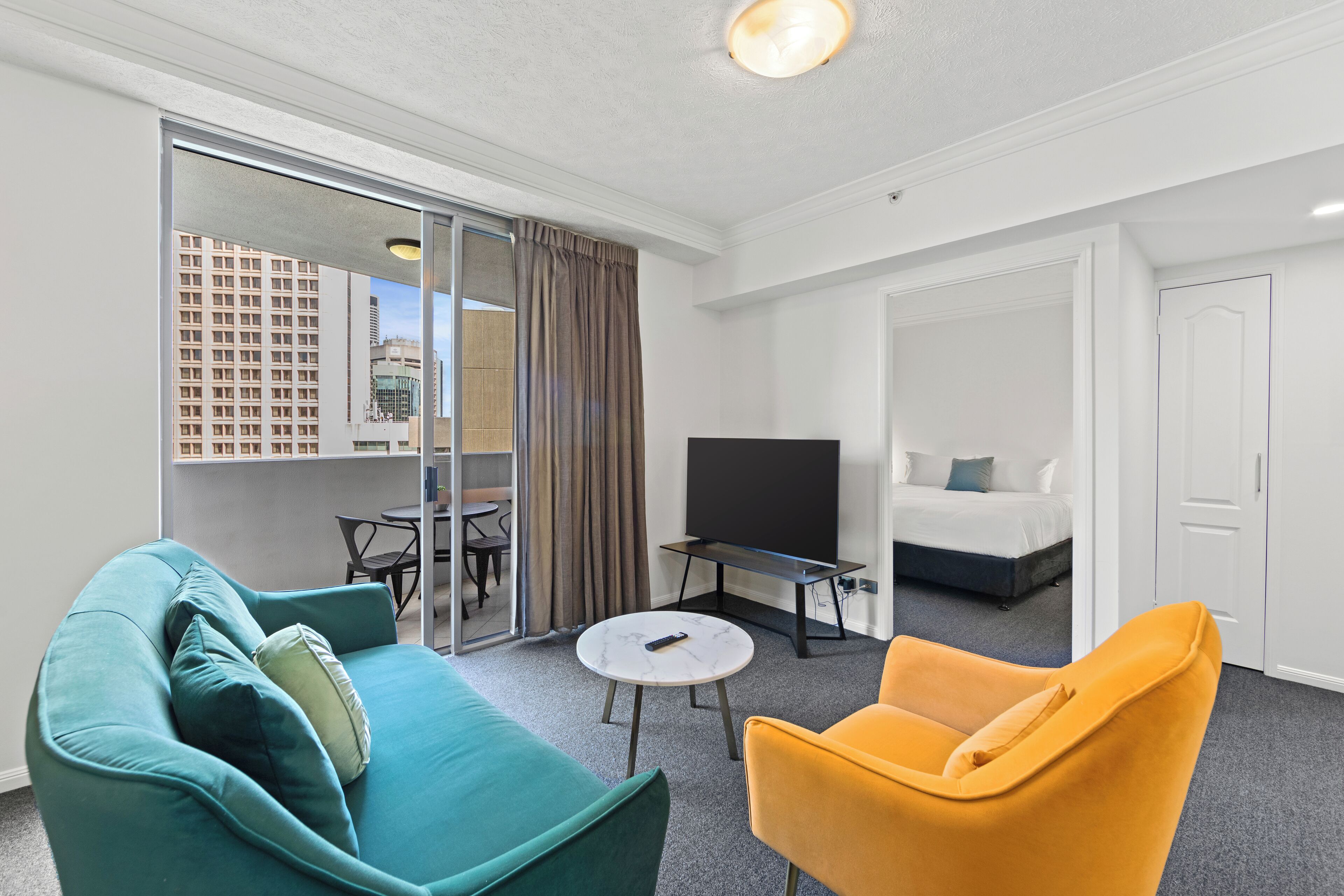 A modern hotel room with a teal sofa, a yellow armchair, a flat-screen TV, a small table, and a sliding glass door leading to a balcony with a city view.