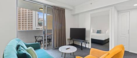 A modern hotel room with a teal sofa, a yellow armchair, a flat-screen TV, a small table, and a sliding glass door leading to a balcony with a city view.