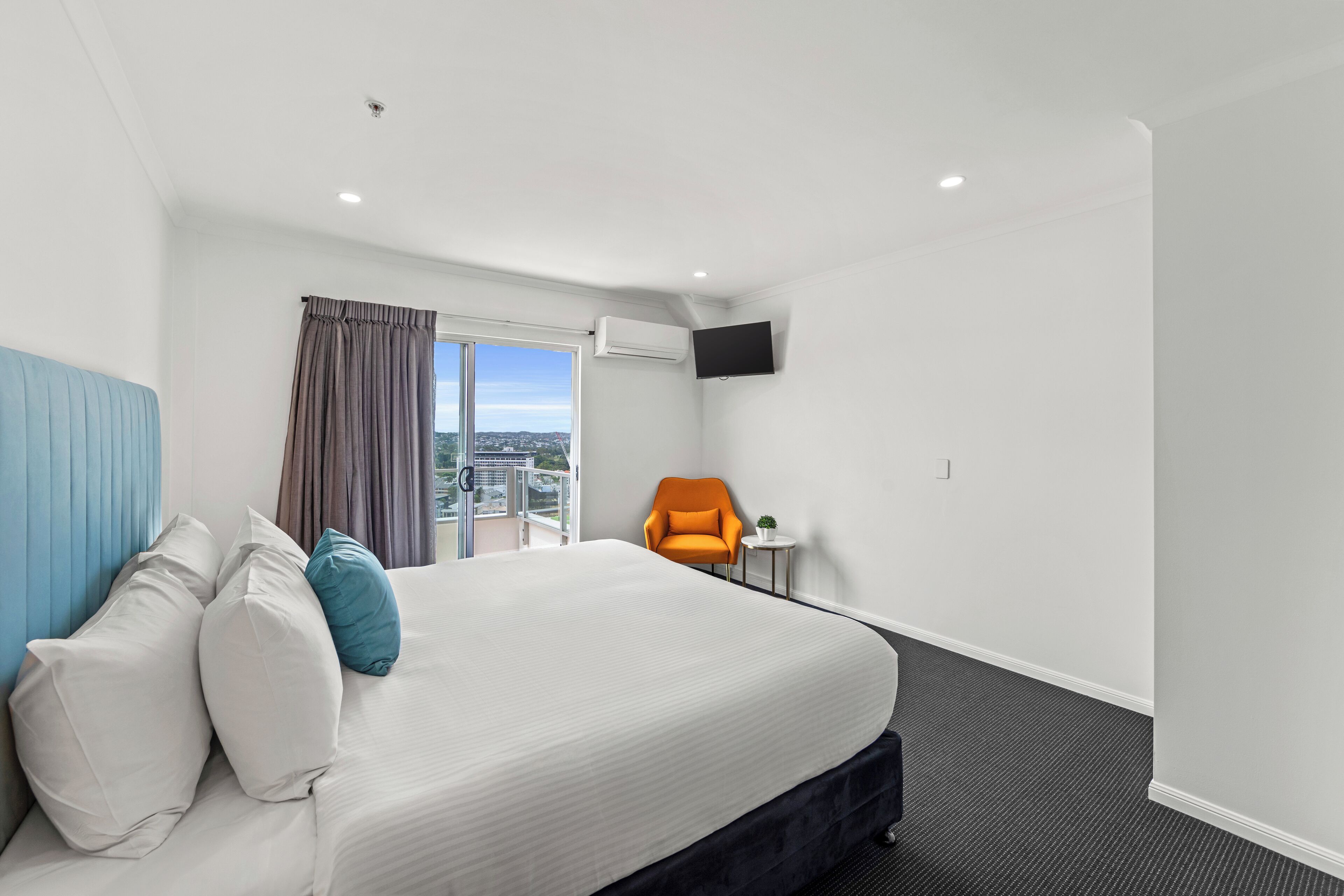 A hotel room with a large bed, a blue headboard, an orange chair, a small table, and a TV mounted on the wall.