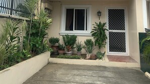 Une maison à deux étages avec un toit rouge, une véranda et un petit jardin où poussent différentes plantes.