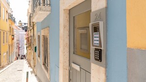 A narrow street with buildings painted in pastel colors. A door with a keypad lock and a number '24' sign is visible. - GuestReady - Esperança do Cardal R/C (Lisbon)