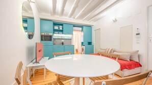 A compact living space with a round dining table, wooden chairs, a sofa with a red blanket, and a kitchen area with teal cabinets and modern appliances. - GuestReady - Esperança do Cardal R/C (Lisbon)