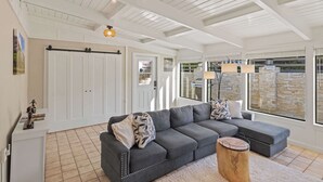 Un vaste salon doté d’un grand canapé d’angle gris, d’une table basse en bois et d’un plancher blanc. Les murs sont blancs, le plafond est orné de poutres apparentes et de grandes fenêtres offrent une vue sur un mur en pierre.