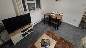 Ein Wohnzimmer mit Fernseher auf einem Schrank, einem Esstisch mit Stühlen, einem Fenster mit Vorhängen, einer Heizung und einem kleinen Teppich.
