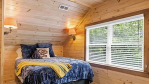 Un dormitorio con techo de madera, un ventanal con persianas, una cama con cubrecama azul, dos lámparas de pared y un asiento junto a la ventana.
