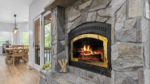 Una chimenea de piedra con fuego, una zona de comedor con mesa y sillas de madera, y vistas al exterior.
