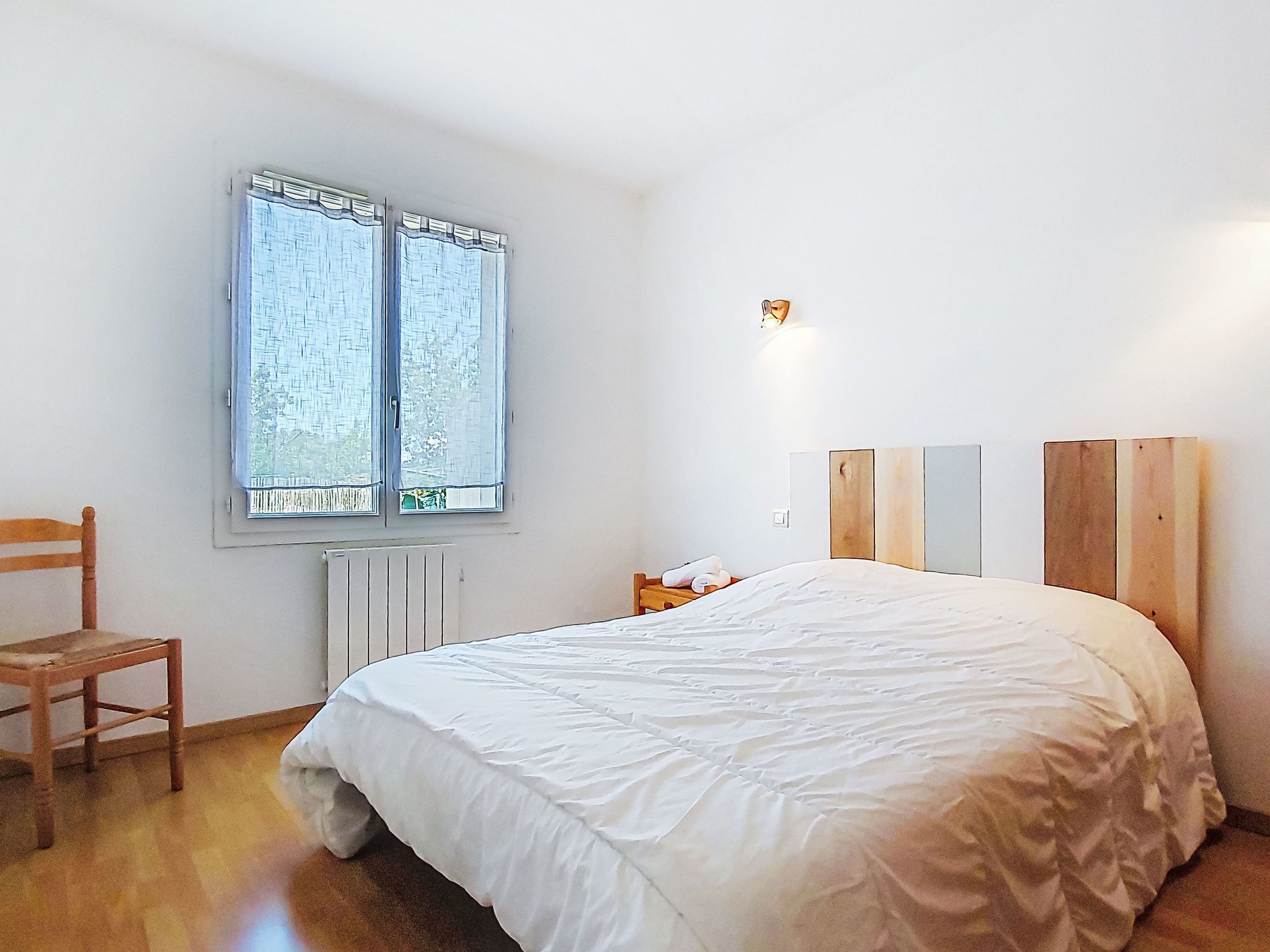 A bedroom with a bed, a chair, a window, and a wall-mounted light fixture.