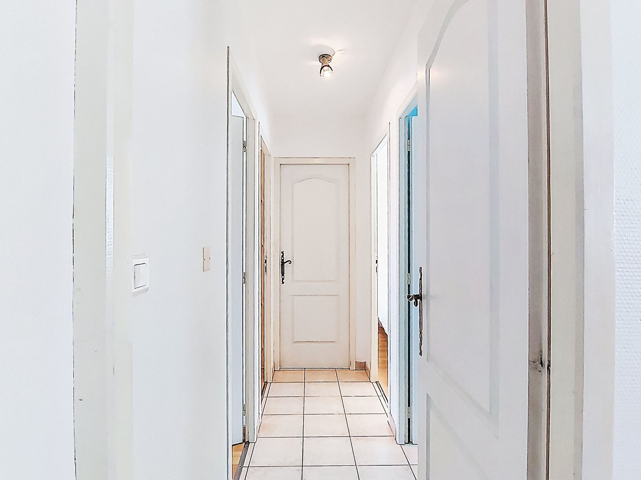 Um corredor estreito com paredes brancas e chão de azulejos, que leva a uma porta no final.