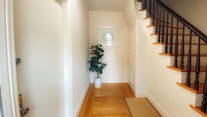 A hallway with a wooden floor, a potted plant, a framed picture on the wall, and a staircase with wooden railings. - Charming 2-bedroom house super close to Brown & RISD, Wayland Square (Providence)