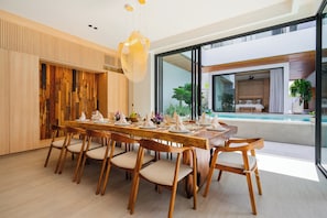 A modern dining area with a wooden table set for six, large windows, and a view of a pool and outdoor space. - Villa Brandon by Nakula (Kedonganan)